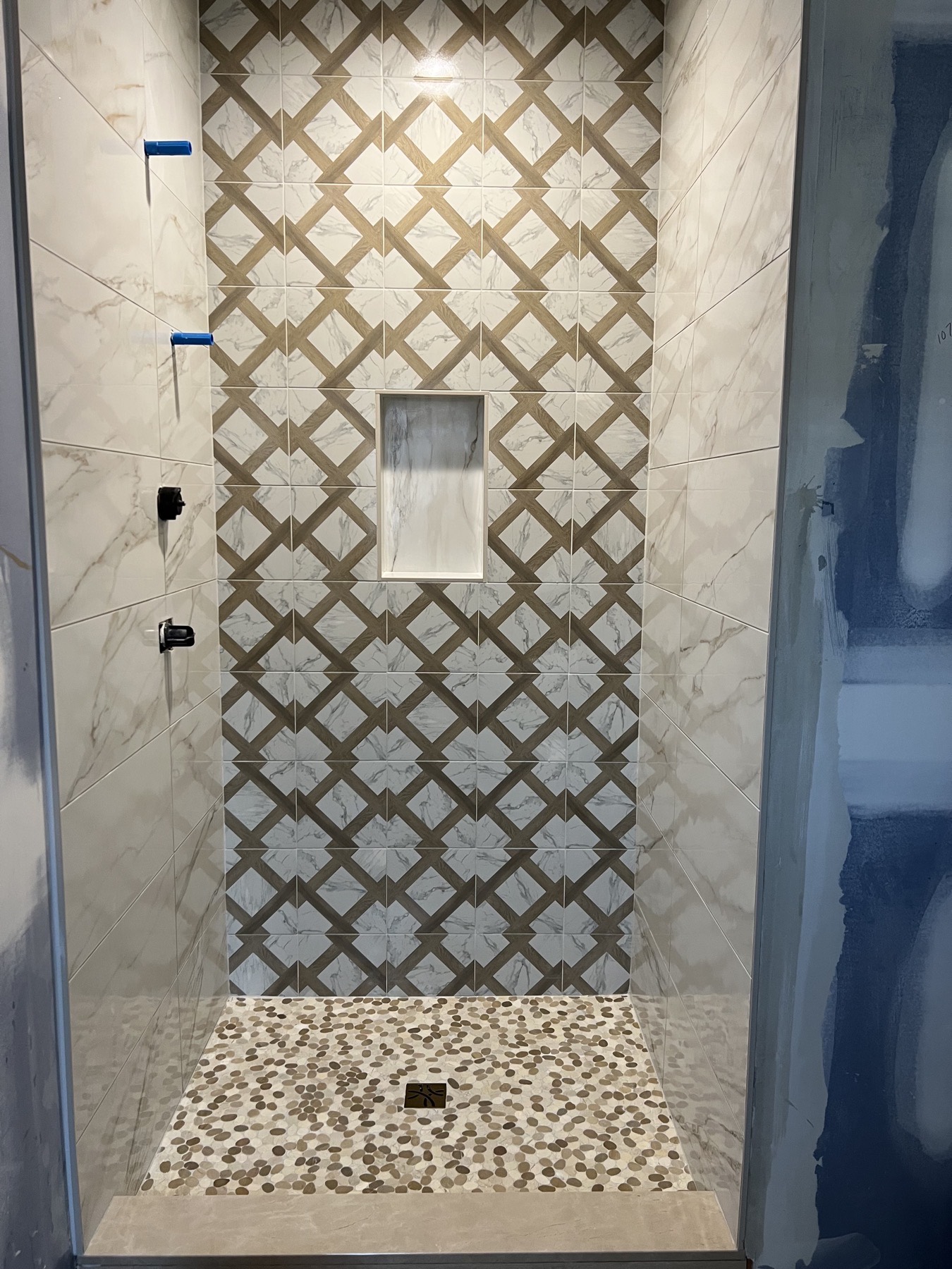 Shower feature wall in Whitehouse Station, NJ built from a woven lattice pattern of wood-look and marble-look tile, above a pebble mosaic shower floor