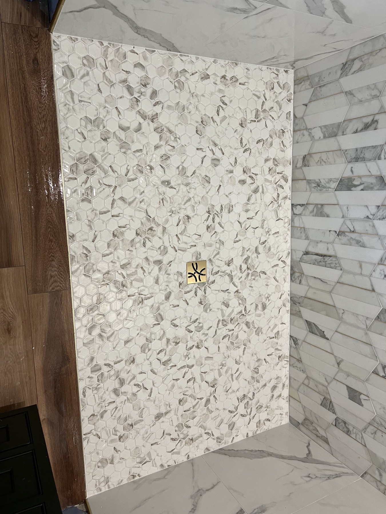 Hexagon marble-look mosaic shower floor in Whitehouse Station, NJ with a square brushed brass drain, meeting wood-look plank tile at a brass transition strip
