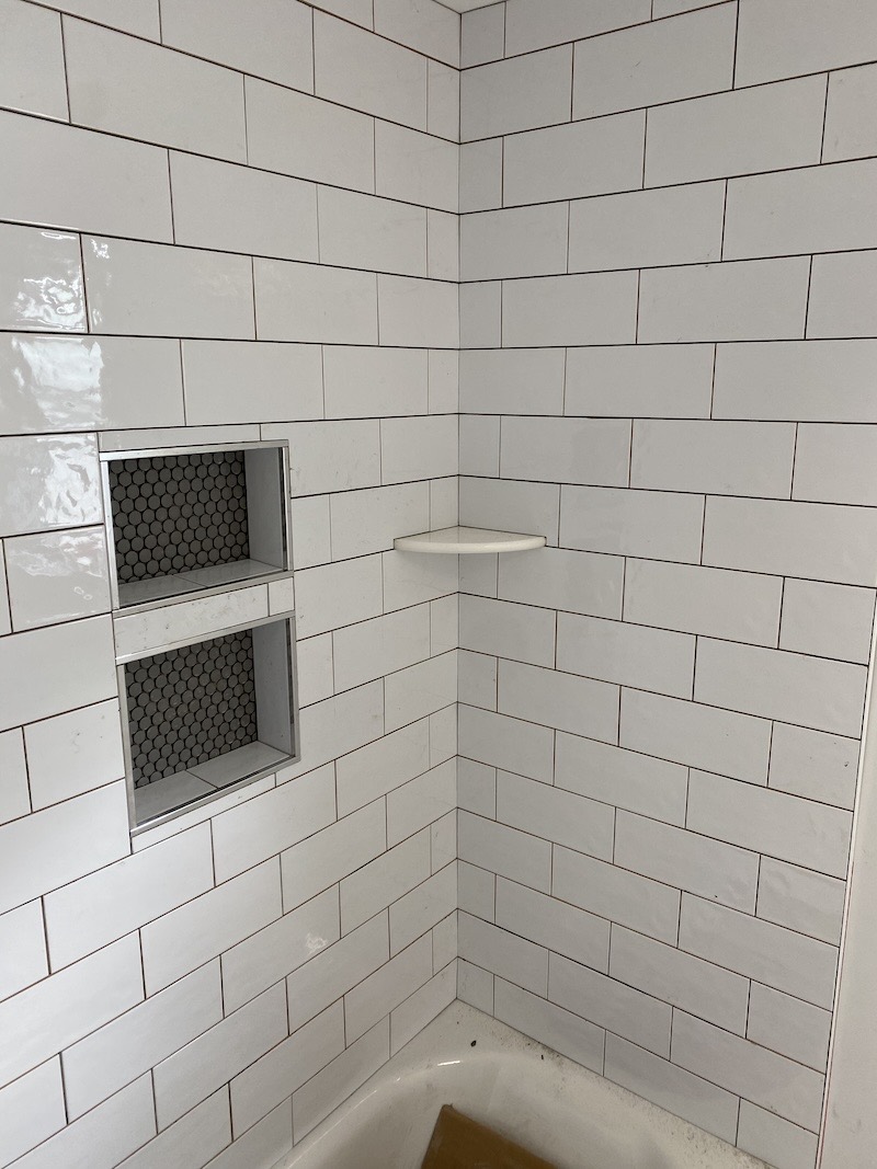 White subway tile tub surround in Somerville, NJ with dark grout, a two-compartment recessed niche backed in black penny round mosaic and a solid corner shelf