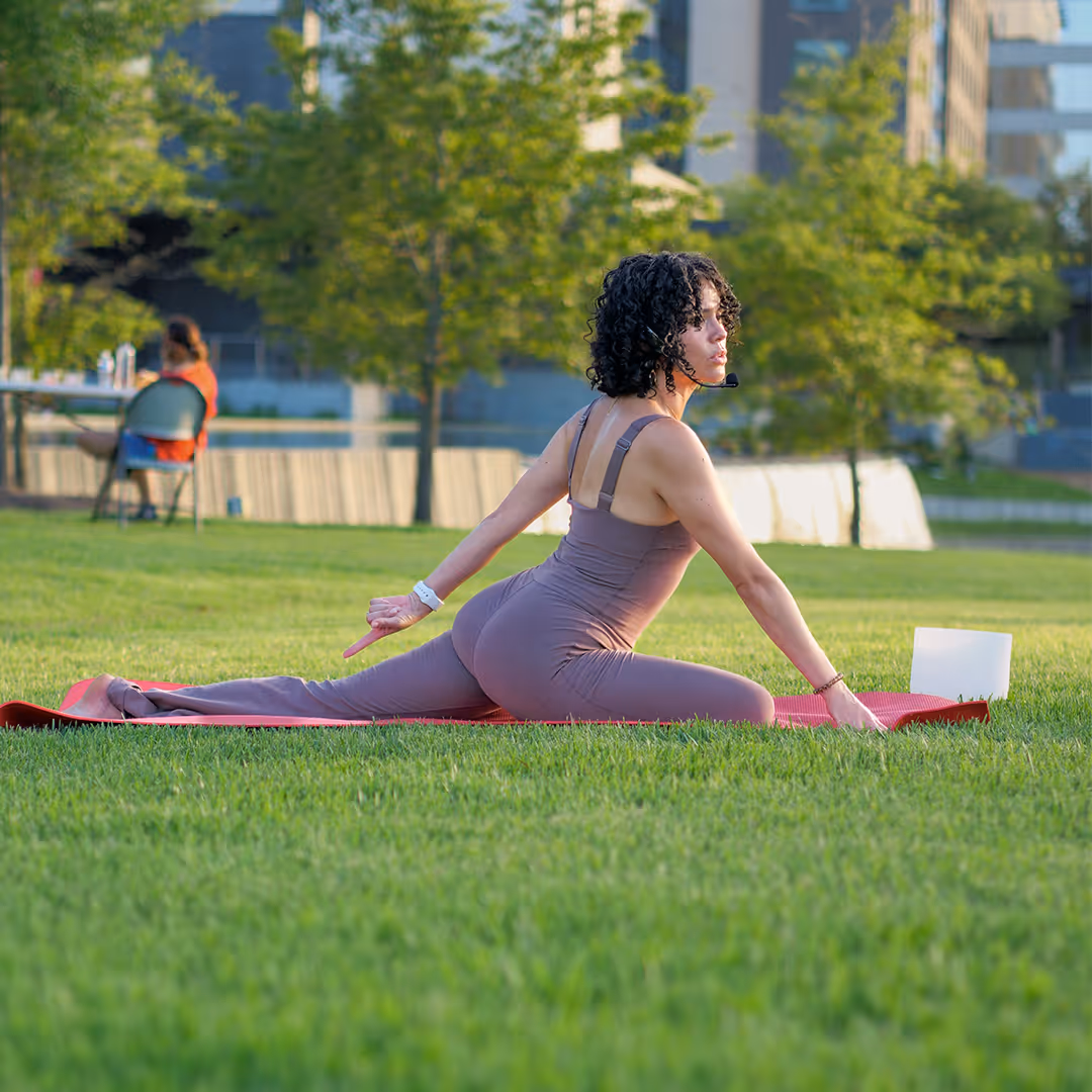 Woman performing yoga stretch on a red mat in a grassy park with trees and buildings in the background.