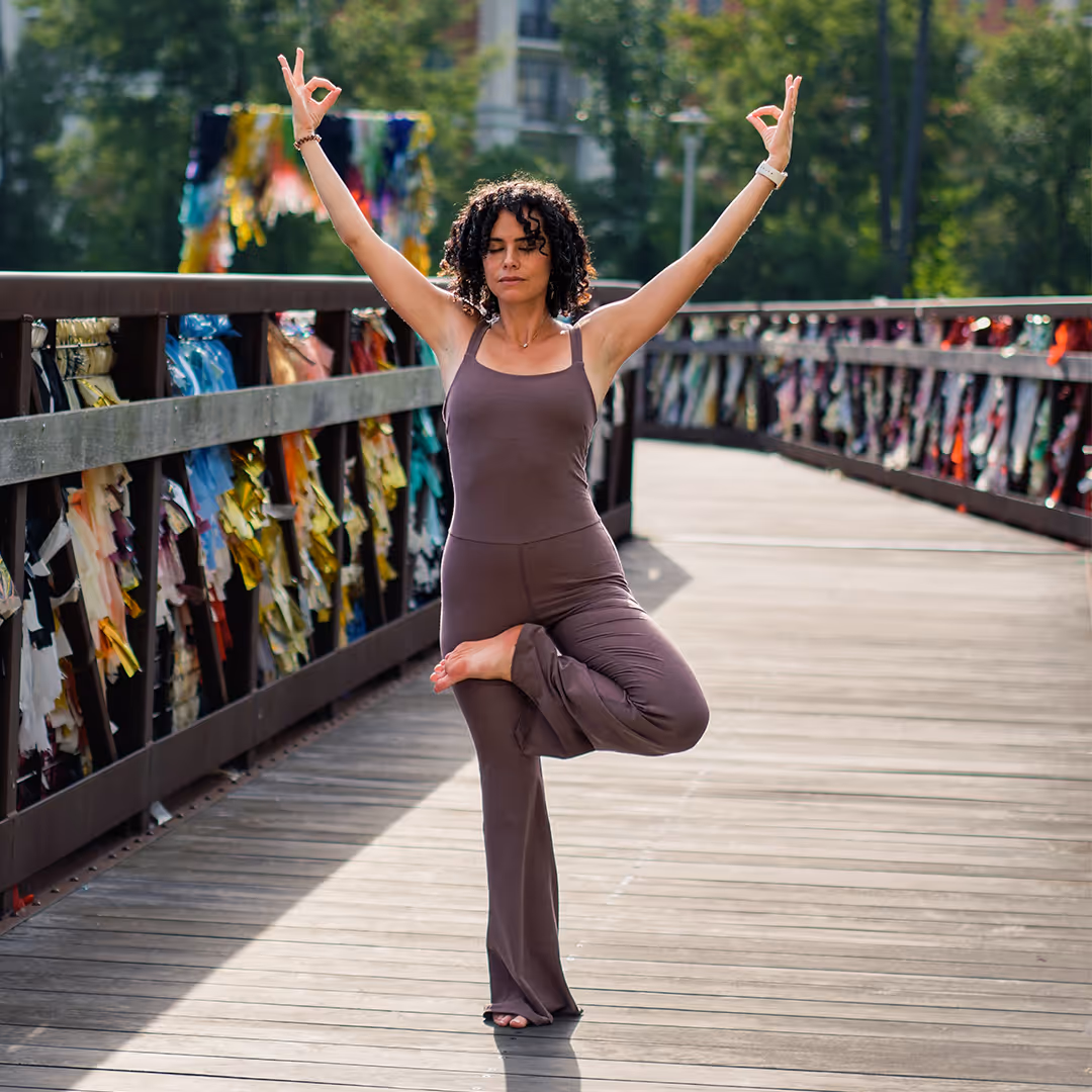 Woman practicing yoga in tree pose with arms raised and eyes closed on a wooden bridge outdoors.
