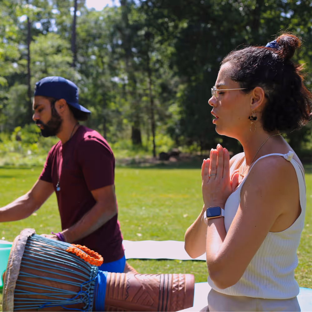 A woman with glasses and a smartwatch sits on grass with hands in prayer position, while a man in a backwards cap plays a drum beside her in a sunlit park.