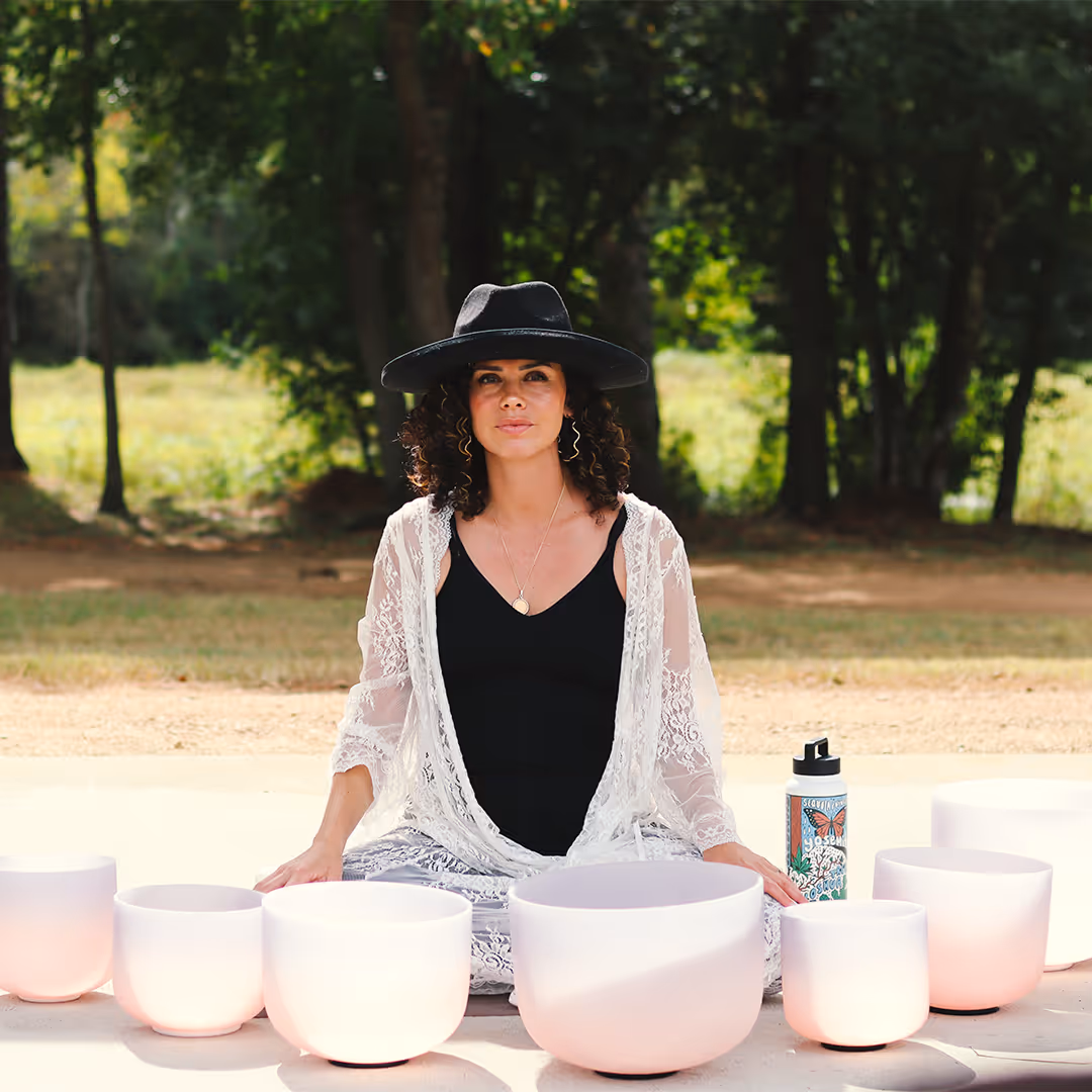 Woman with curly hair wearing a black hat and white lace shawl sits cross-legged outdoors surrounded by white crystal singing bowls.