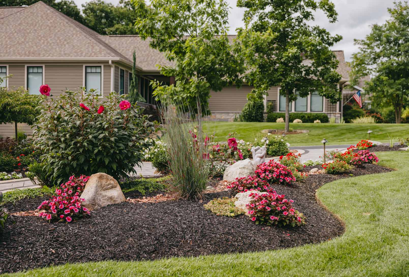 Front yard flower garden with red blooms, stone boulders, and manicured grass in a Chicago suburban neighborhood.