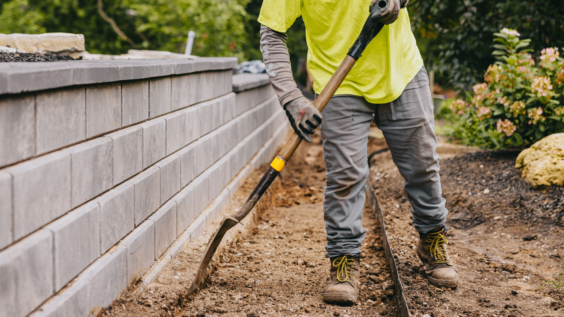 Close-up of landscaper shaping dirt path beside retaining wall construction.