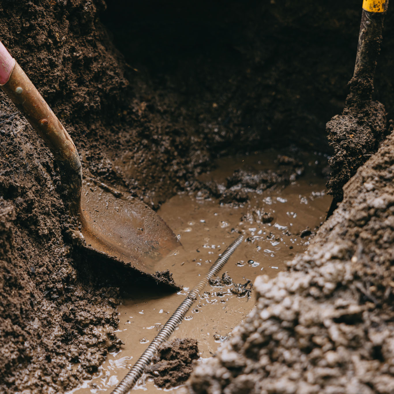 Shovel digging through muddy soil with drainage pipe visible during a landscape installation in the Chicago suburbs.