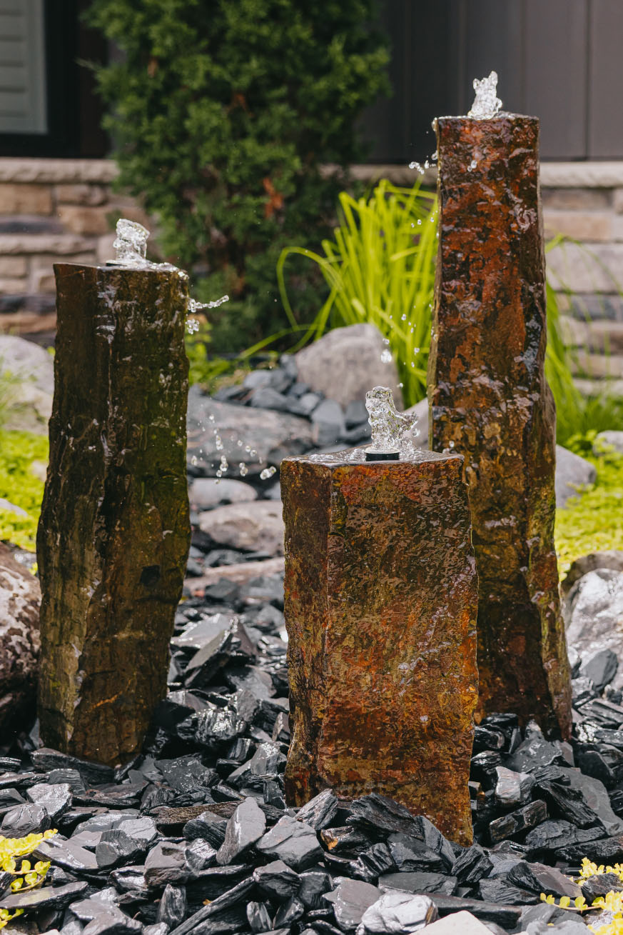 Three stone water fountains with bubbling water feature surrounded by dark river rock landscaping