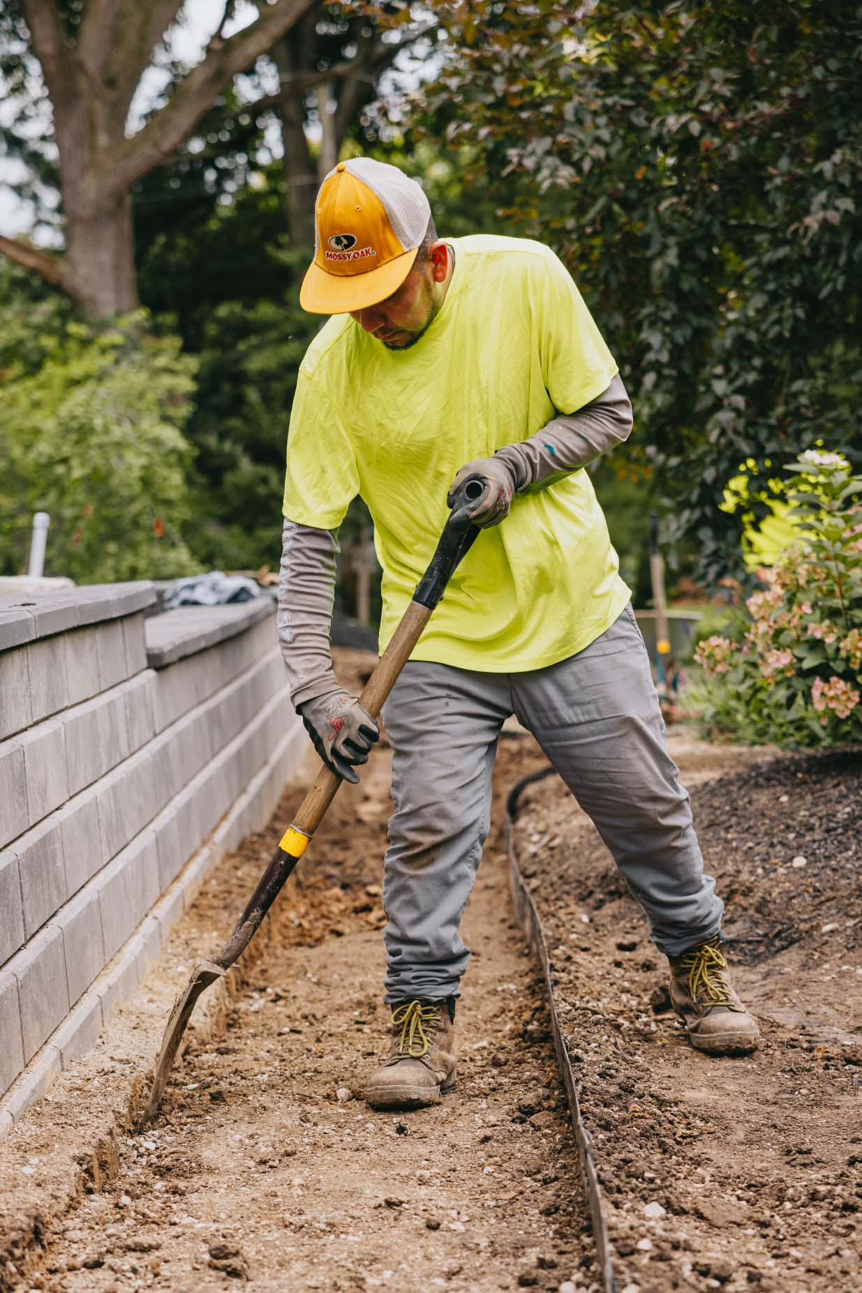 Landscaping crew member digging soil to prepare a pathway foundation beside a retaining wall