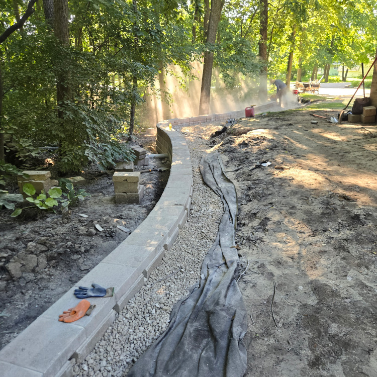 Gravel base and retaining wall under construction with sunlight filtering through trees.