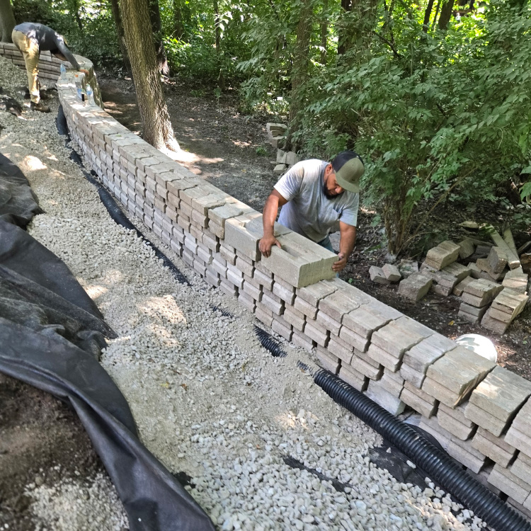 Workers stacking stone blocks to build a retaining wall surrounded by wooded terrain.