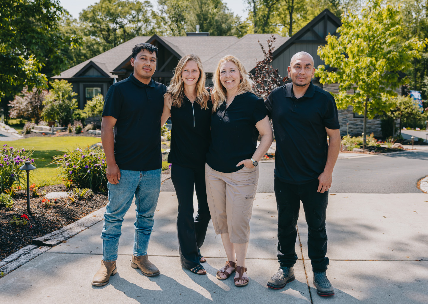 Four people standing on a driveway outside a house, smiling, with lush landscaping around them.