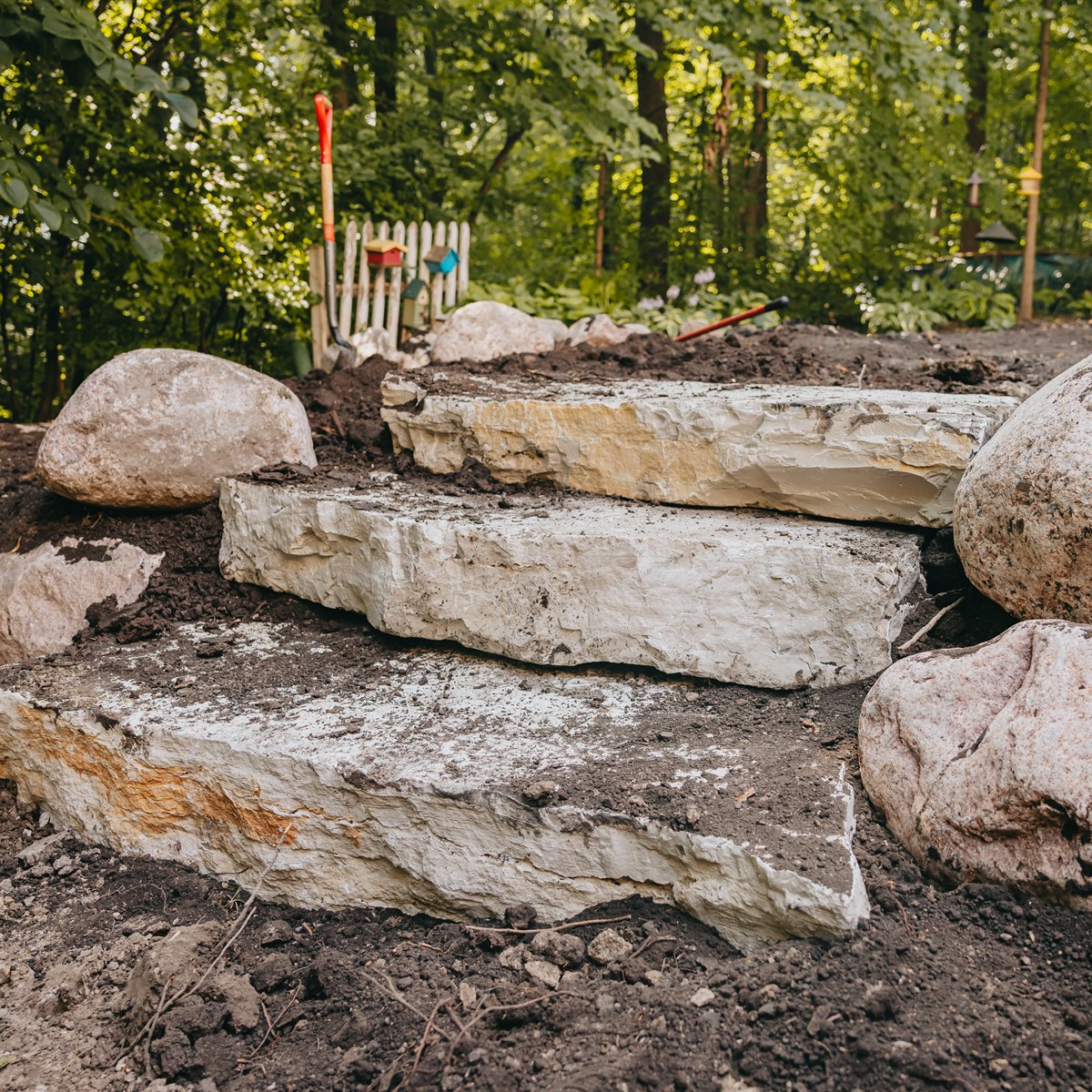 Three large, rough-hewn stone steps stacked outdoors in a garden setting with dirt and large boulders surrounding them, and garden tools and greenery in the background.