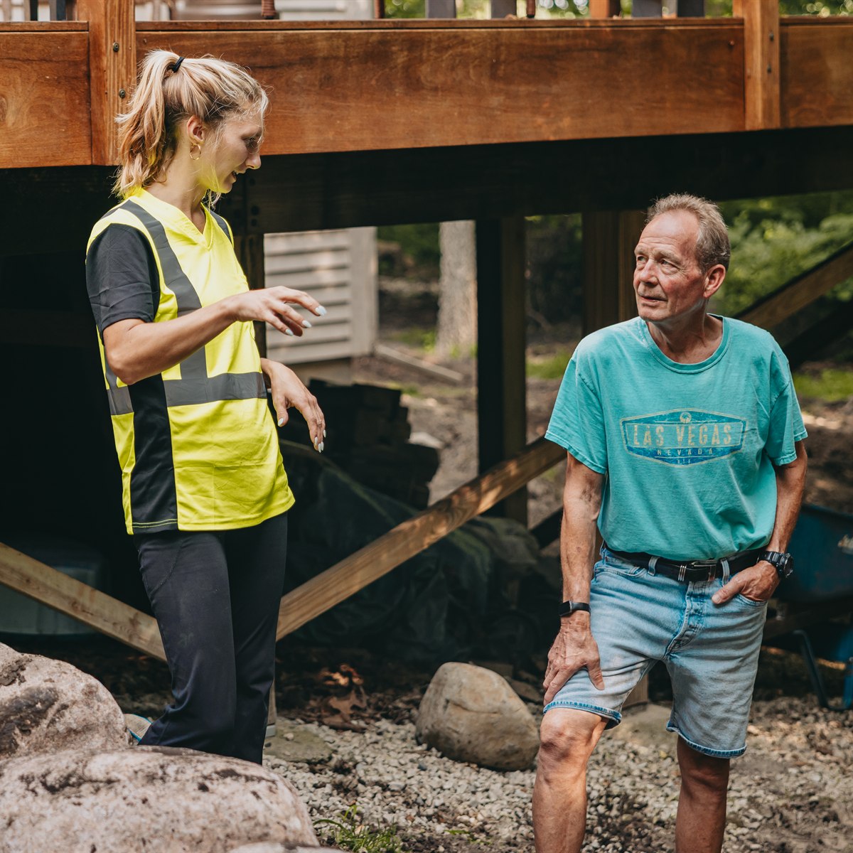 A woman in a yellow safety vest is talking to an older man wearing a turquoise Las Vegas t-shirt and denim shorts outdoors near a wooden deck structure.