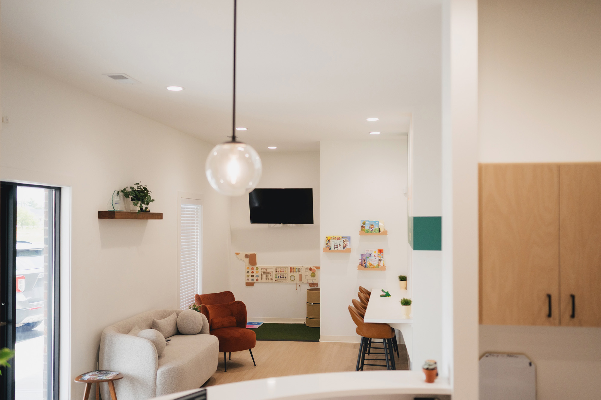 Modern waiting area with couch, chair, wall-mounted TV, stools, and children's books on shelves.