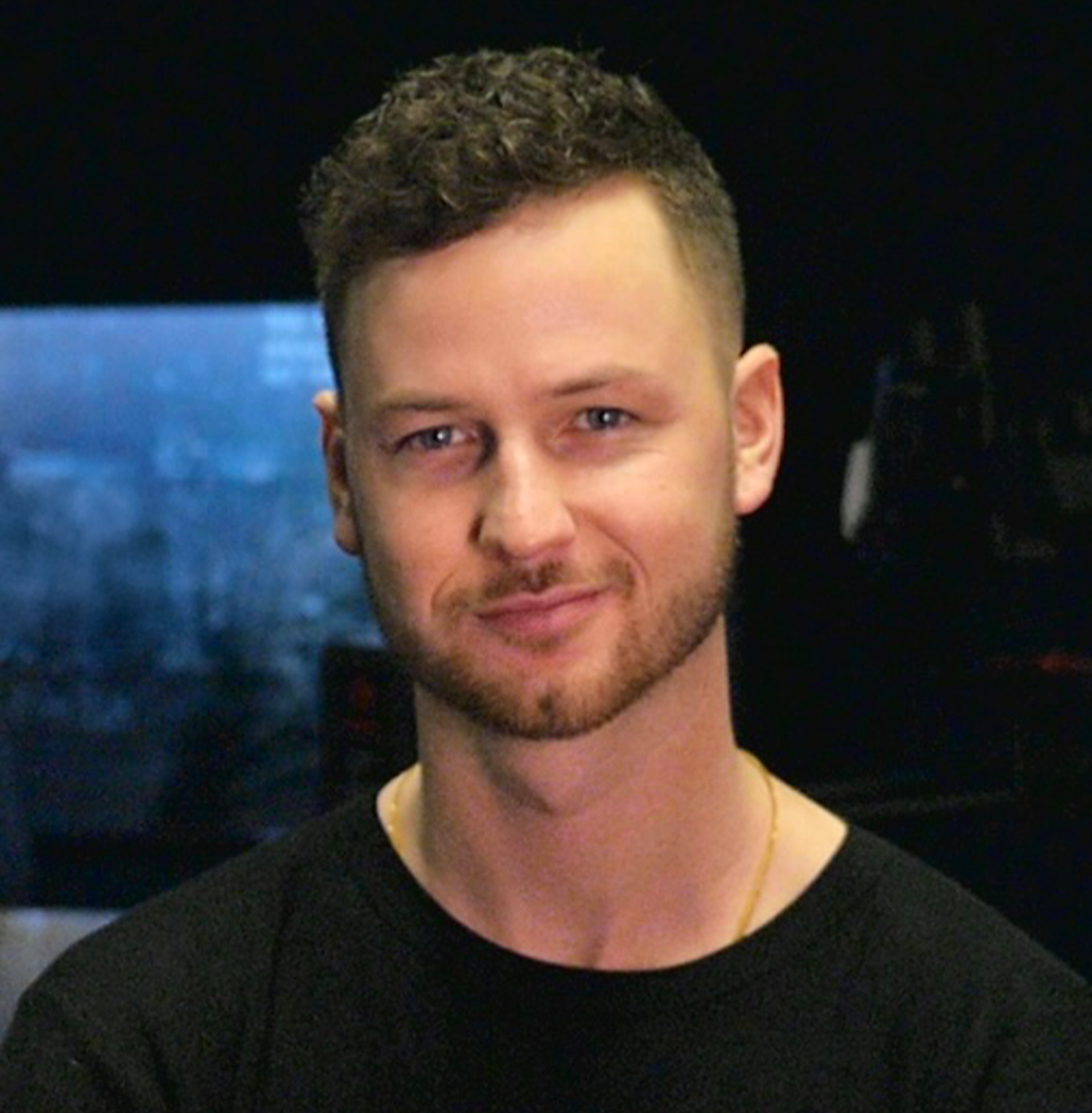 Young man with curly hair and beard smiling, wearing a black shirt and gold necklace against a dark background.