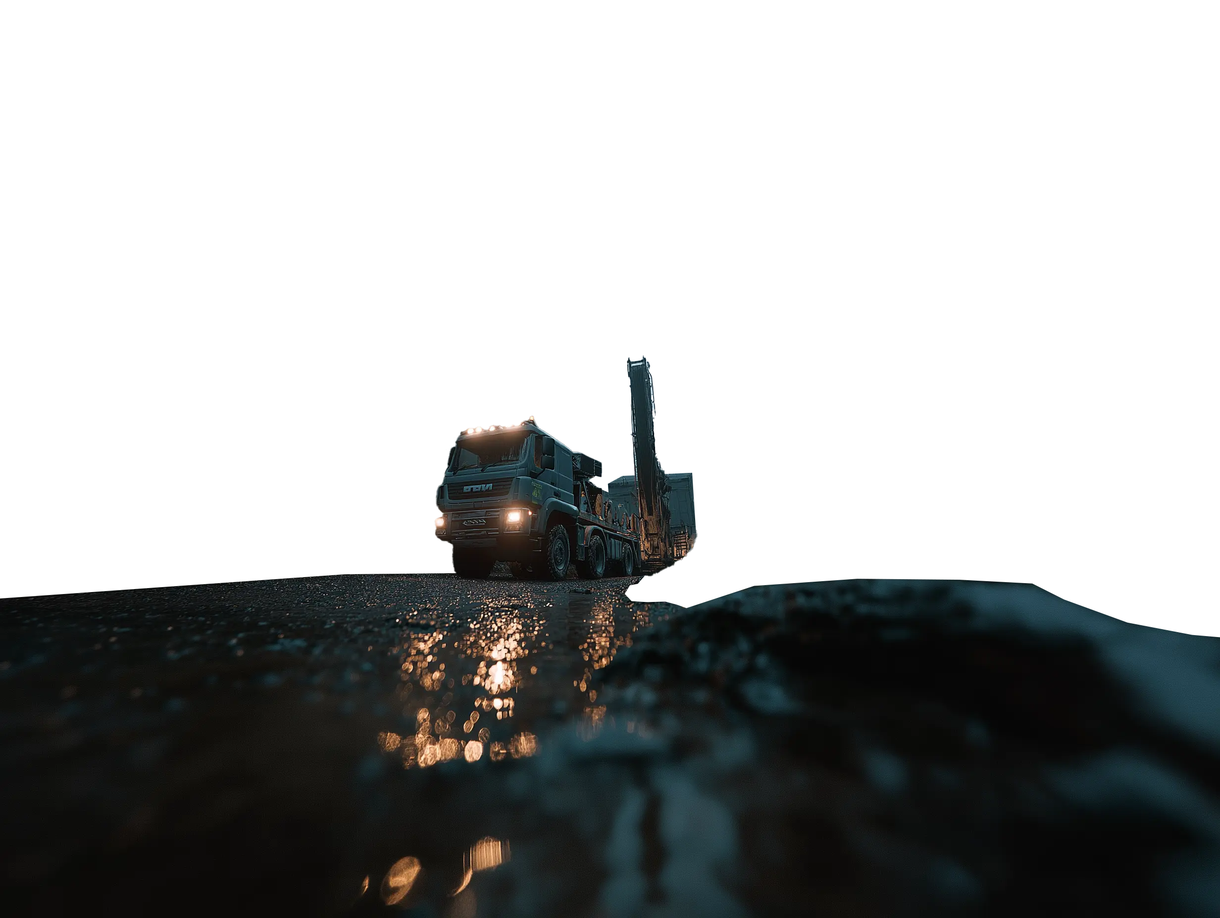 Large drilling truck with headlights on, parked on wet ground reflecting light during early morning or late evening.