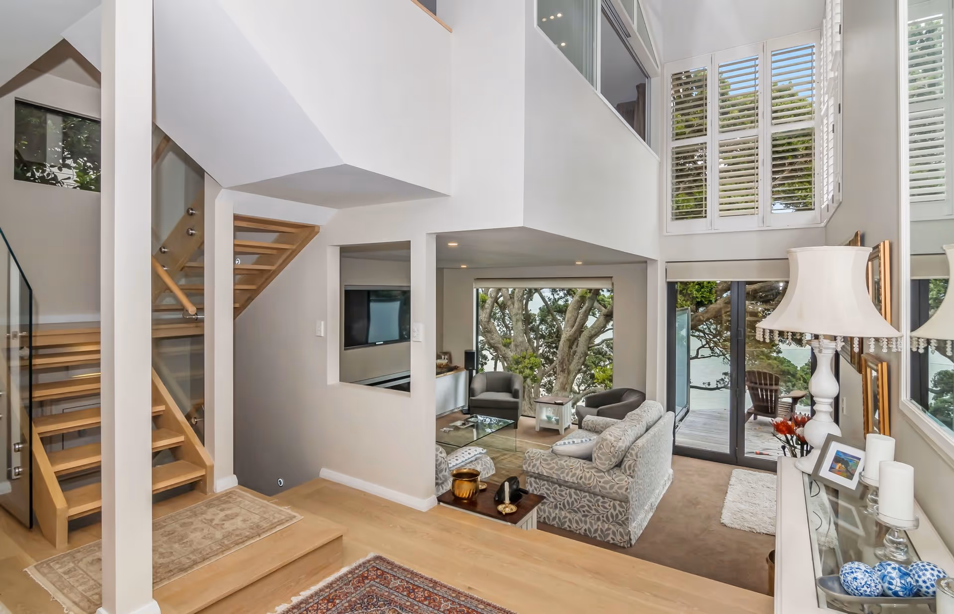 Bright, modern living space featuring a cozy seating area, wooden stairs, and large windows with a view of trees and water outside.