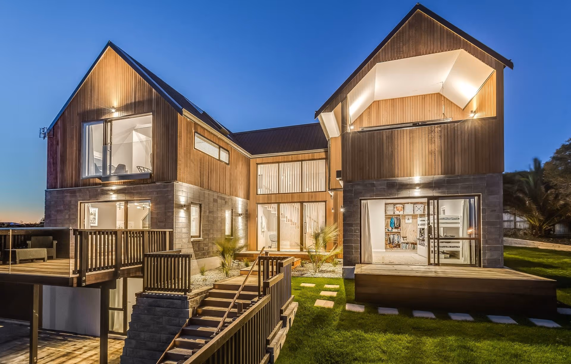 Modern wooden house with large windows and a deck, illuminated at dusk, showcasing a sleek design and lush green surroundings.
