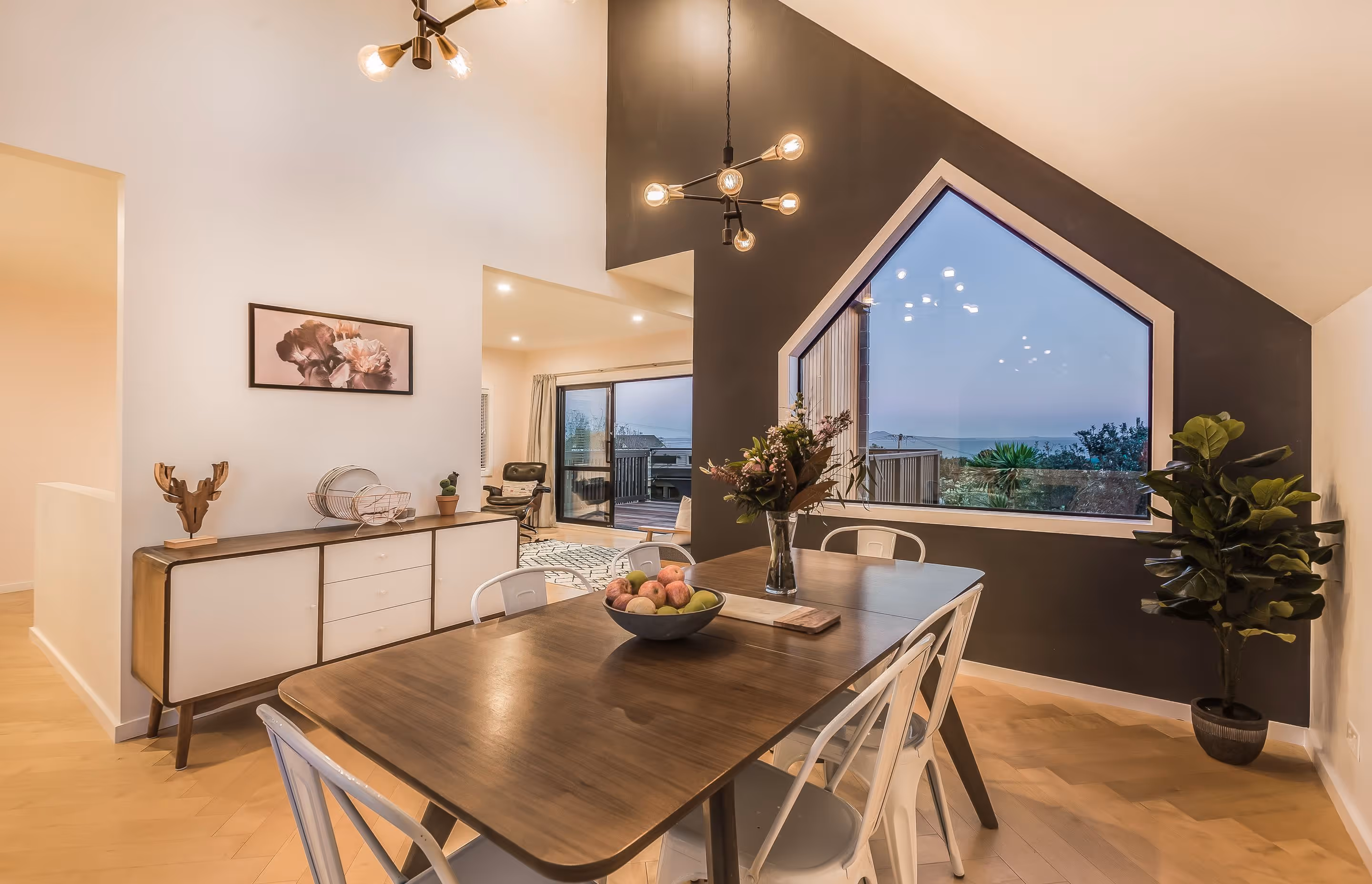 Stylish dining area with a wooden table, white chairs, and a large window showcasing a scenic view at dusk.