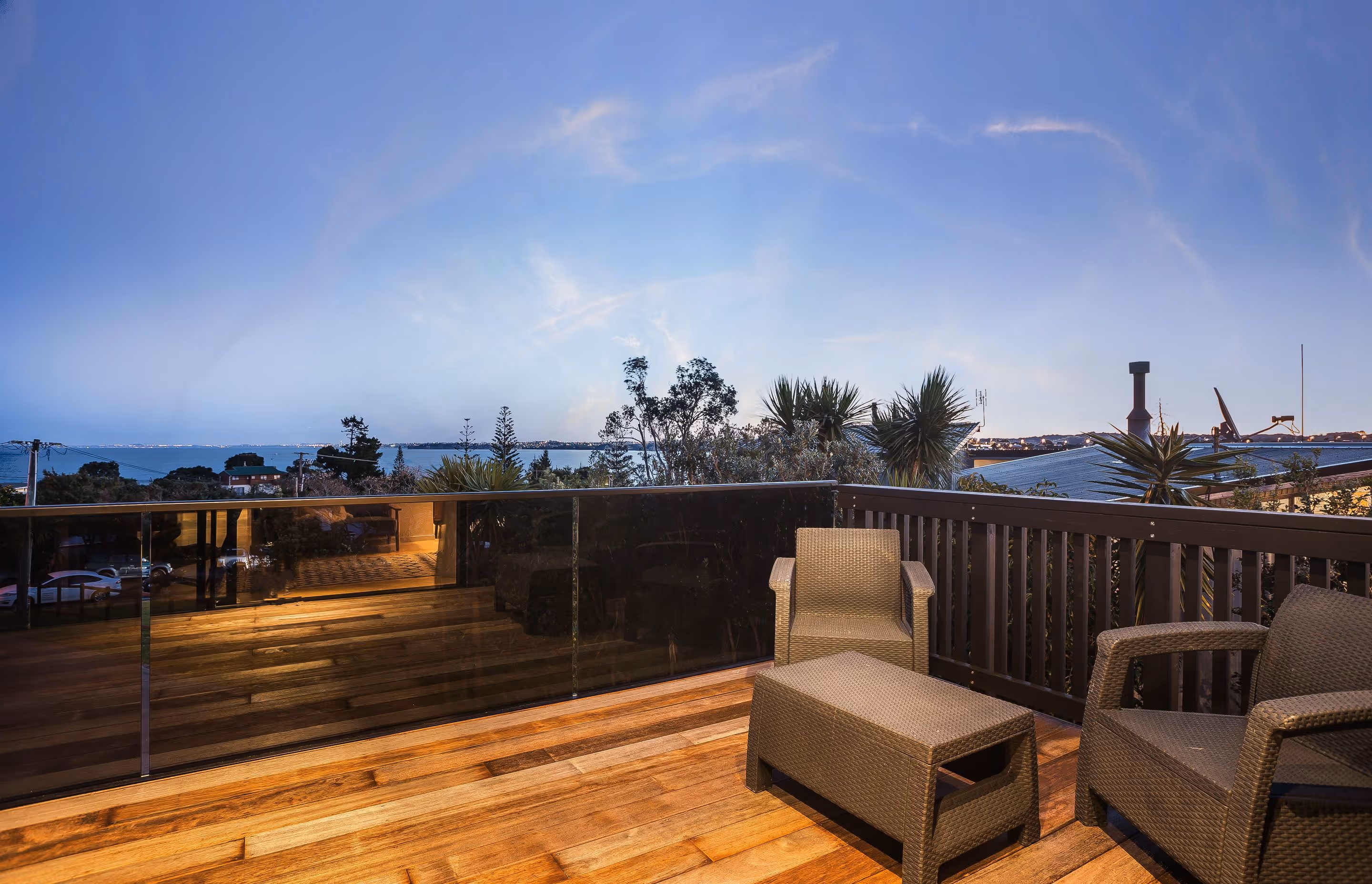 A serene balcony with wooden flooring and wicker chairs, overlooking a peaceful view of trees and a distant seascape at twilight.