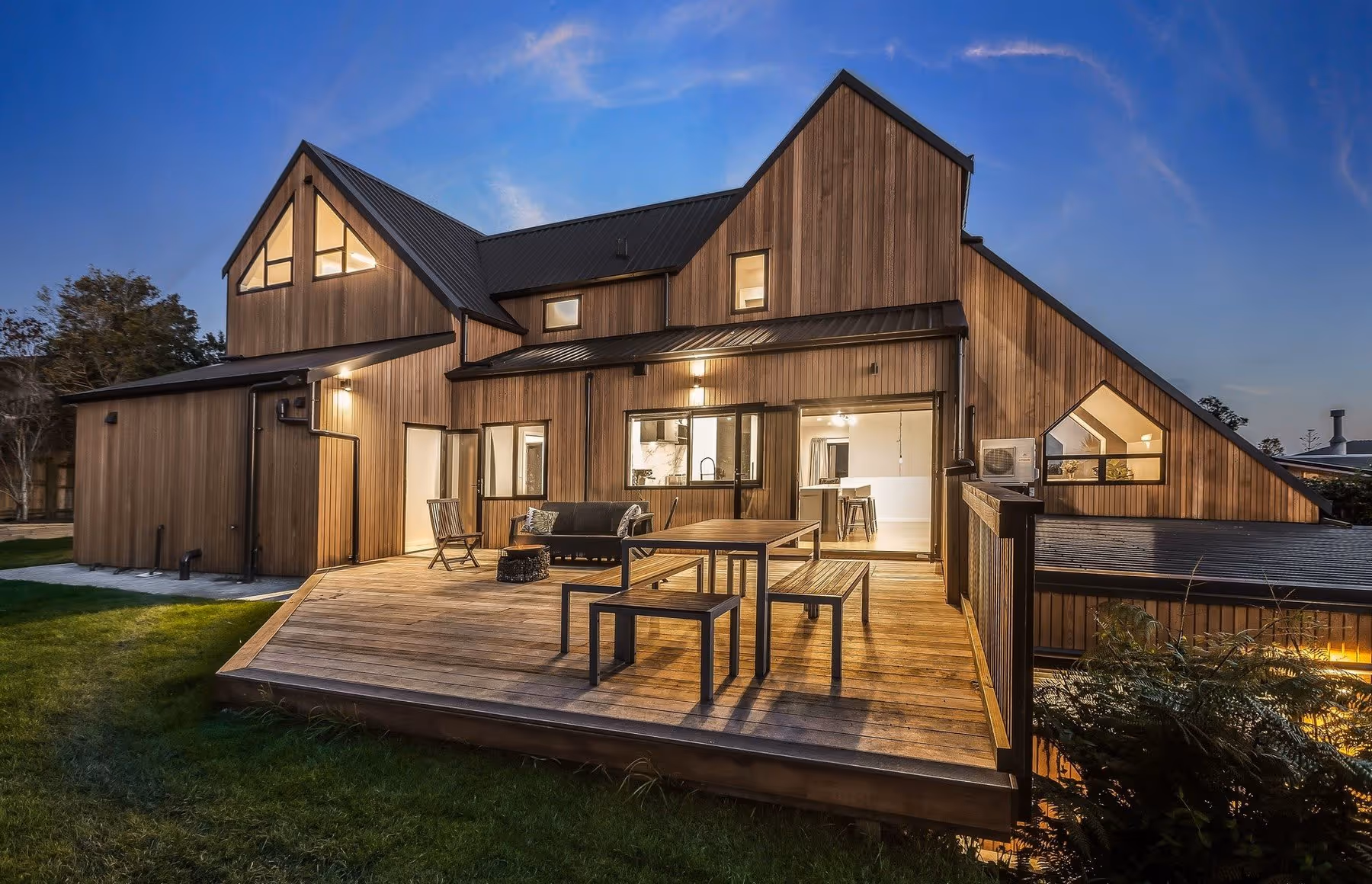 Modern wooden house with a spacious deck, cozy seating, and large windows, illuminated at dusk against a clear blue sky.