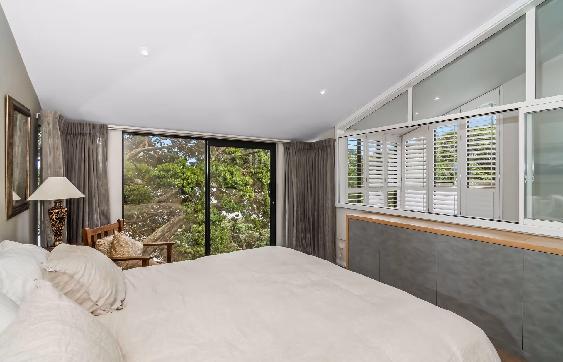 Cozy bedroom featuring a large window with greenery view, light curtains, a comfortable bed, and modern decor.