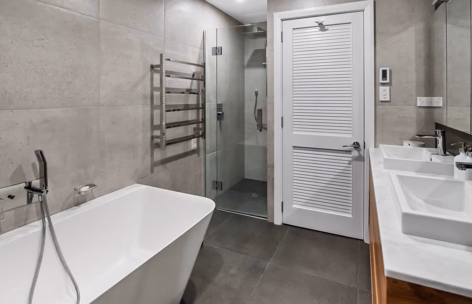 Modern bathroom featuring a white freestanding tub, glass shower, double sinks, and sleek gray tile walls. Elegant and spacious design.