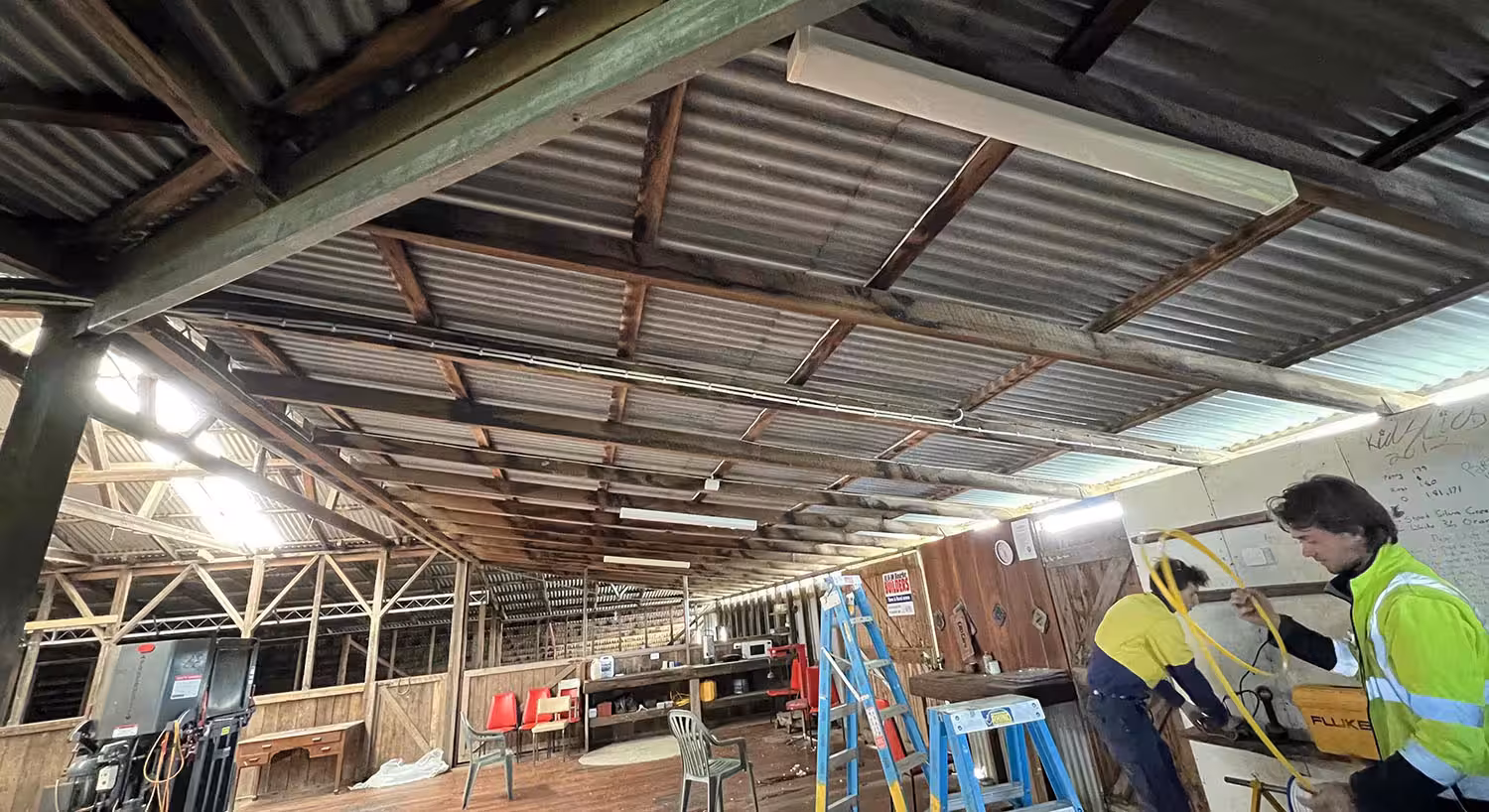 Two men in work attire fix a rustic ceiling inside a large, cluttered workshop. Ladders and chairs are scattered, creating a busy, industrious atmosphere.
