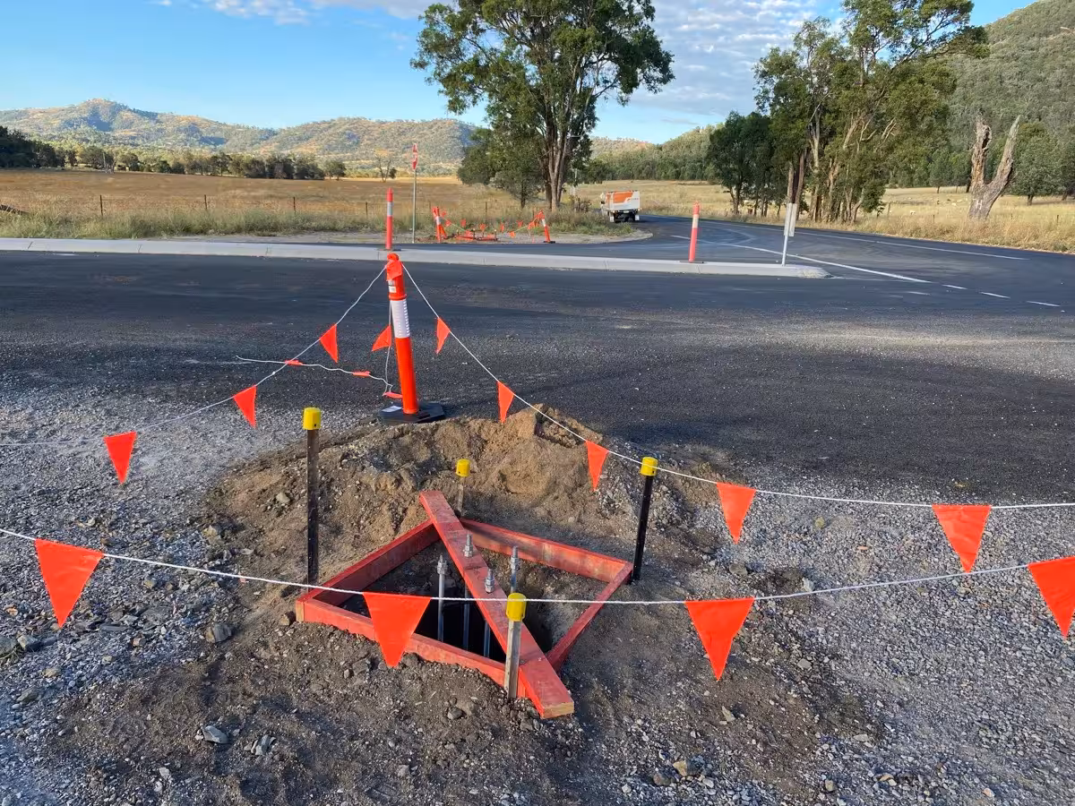 Open triangular metal construction set in the ground surrounded by red and white safety flags at a road intersection under clear sky.