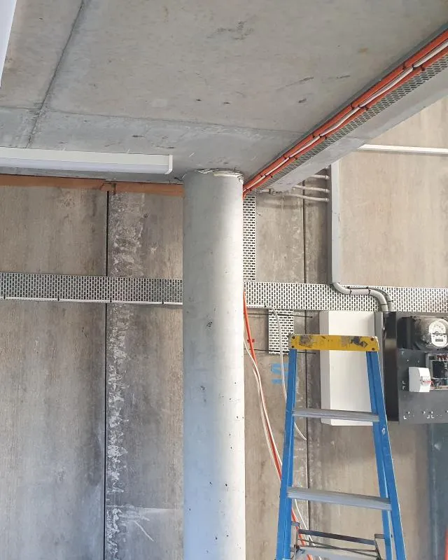 Concrete column in an unfinished room with exposed pipes and cables, next to a blue and yellow step ladder.