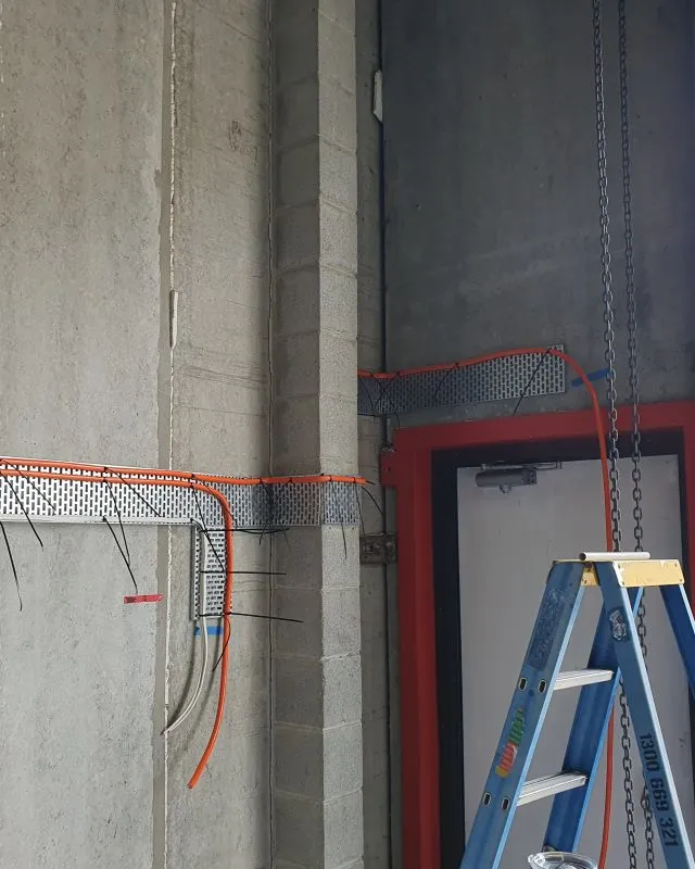 Interior corner of an unfinished room with concrete walls, a closed white door with red trim, blue stepladder, and orange electrical conduit installed along perforated metal supports.