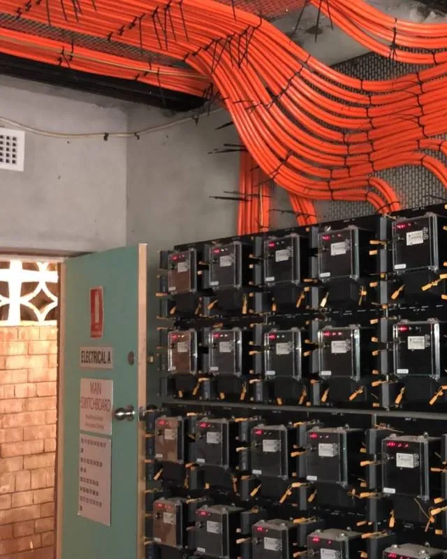 Room with multiple electricity meters mounted on racks and numerous bright orange electrical conduits running overhead.