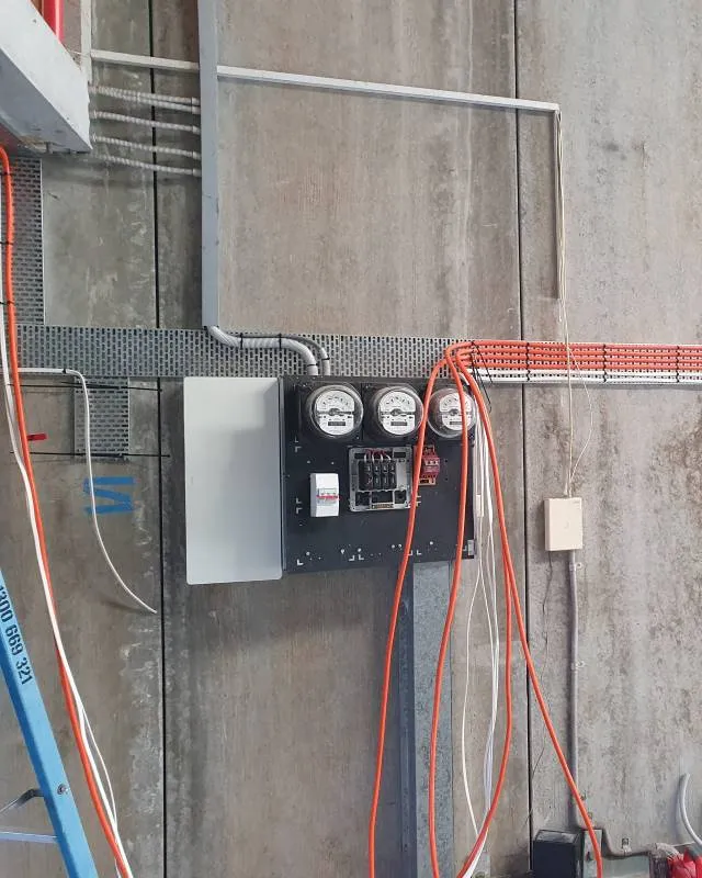 Electric meter panel mounted on concrete wall with three meters and several exposed orange and white electrical cables hanging and running along the wall.