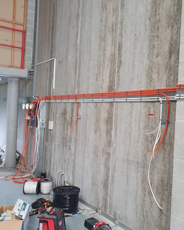 Construction site with exposed gray concrete walls, orange and white electrical conduits installed horizontally, and tools and cable spools on the floor.