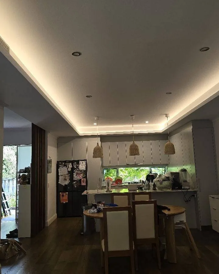 Modern kitchen and dining area with wood flooring, white cabinets, black refrigerator covered with papers, wooden dining table with four chairs, and pendant lights above the kitchen island.