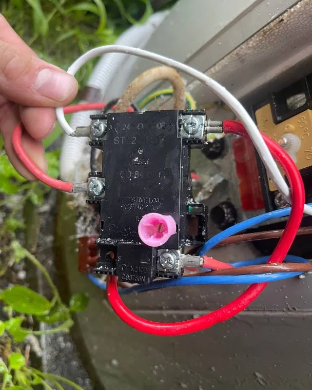 Hand holding a black electrical relay with red, white, blue, and brown wires connected inside an outdoor unit.