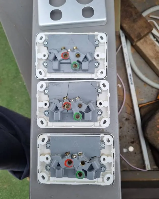 Three gray electrical outlets without faceplates arranged vertically on a metal surface with screws and colored terminal markings in red and green.