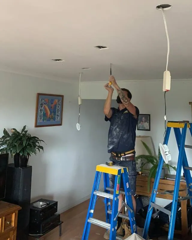 Man wearing a face mask standing on a blue step ladder, using a screwdriver to work on ceiling wiring with multiple hanging cables in a living room.