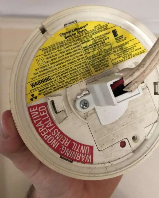 Hand holding a Clipsal Lifesaver smoke alarm with yellow warning label and a red caution label stating 'Warning: Inoperative Until Reinstalled'.