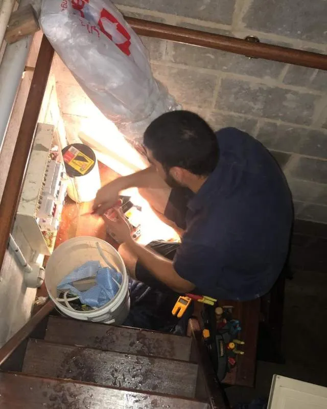 Man in dark shirt repairing an electrical panel on a basement wall, surrounded by tools and a white bucket.