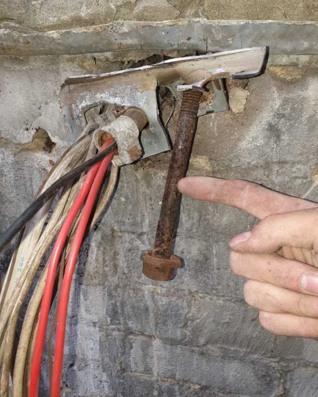 Person's hand pointing at a rusty bolt anchored in a concrete wall with multiple electrical cables nearby.
