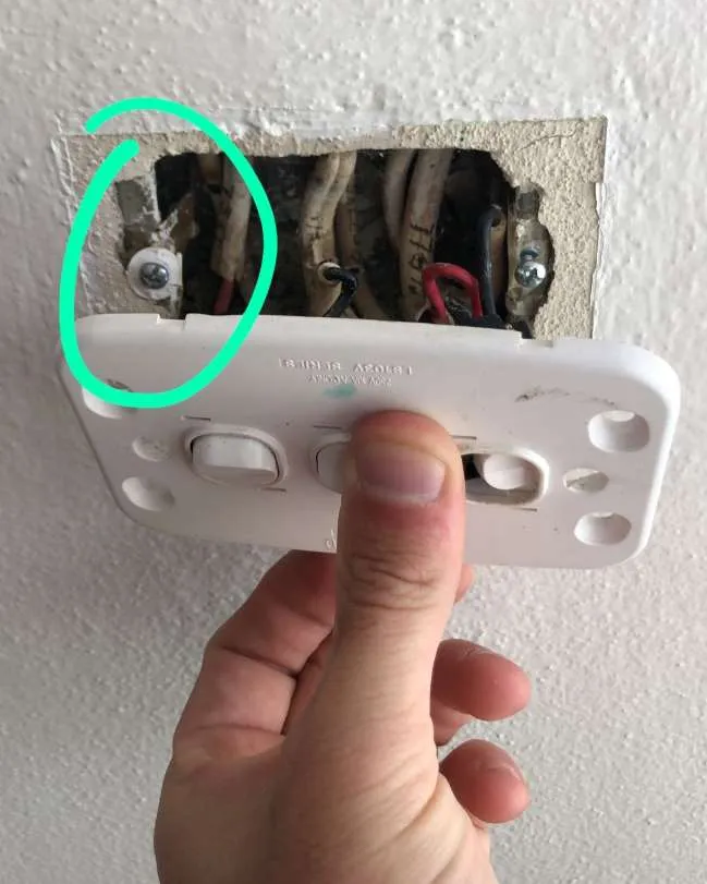 Hand holding a loose light switch cover plate revealing exposed wiring inside the wall with a green circle marking a screw hole on the left side.