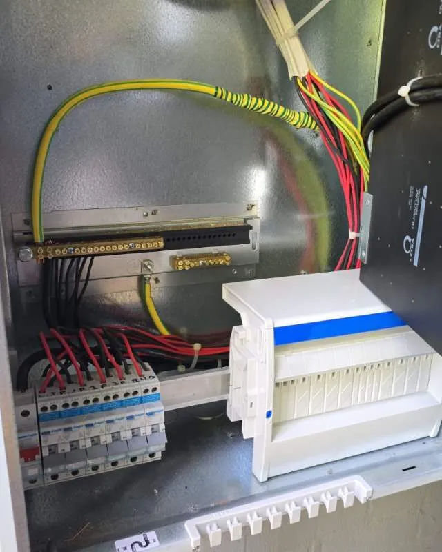 Electrical panel featuring circuit breakers with red and black wires, yellow and green grounding wires, and a white terminal block inside a metal enclosure.