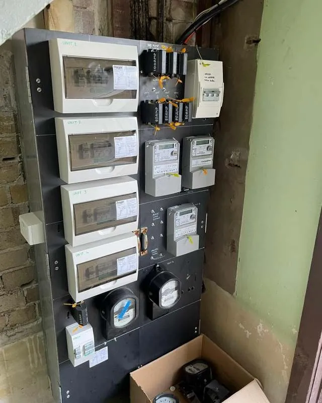 Electric meter panel with multiple unit meters, circuit breakers, and an open cardboard box containing additional meters, mounted on a wall.
