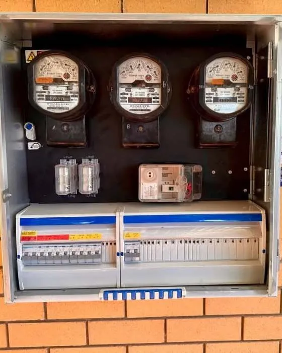 Electrical panel box mounted on a brick wall containing three analog electricity meters and multiple circuit breakers.