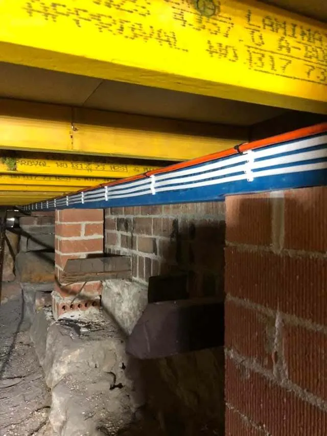 View under a building showing yellow steel beams, brick supports, and a blue and red cable tray running along the length.
