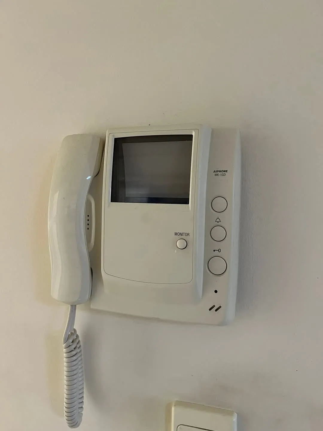 Wall-mounted white Aiphone MK-1GD video intercom with handset and monitor screen.