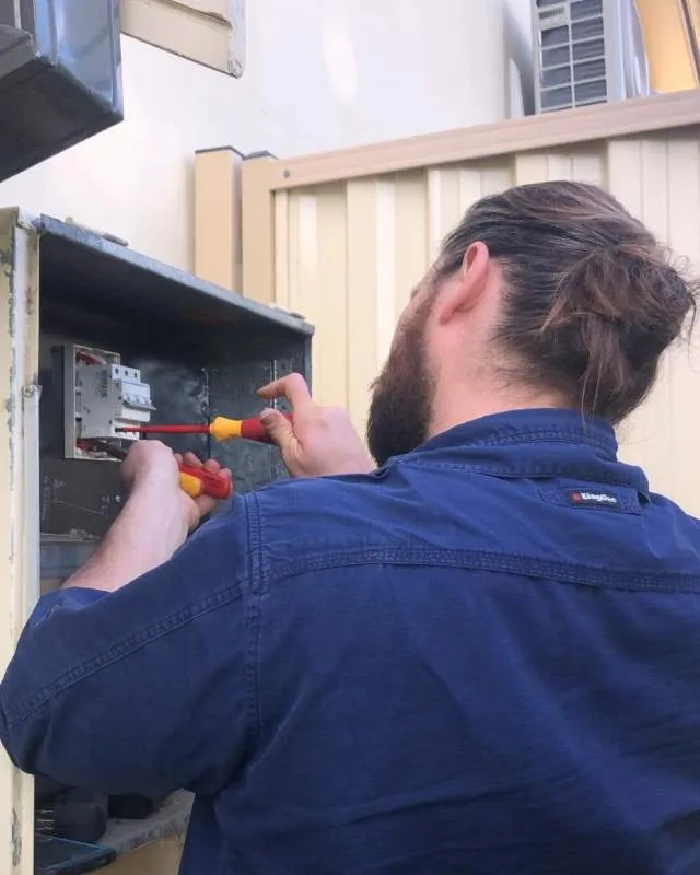 Man with beard and hair tied back using screwdrivers to work on an open electrical panel outdoors.