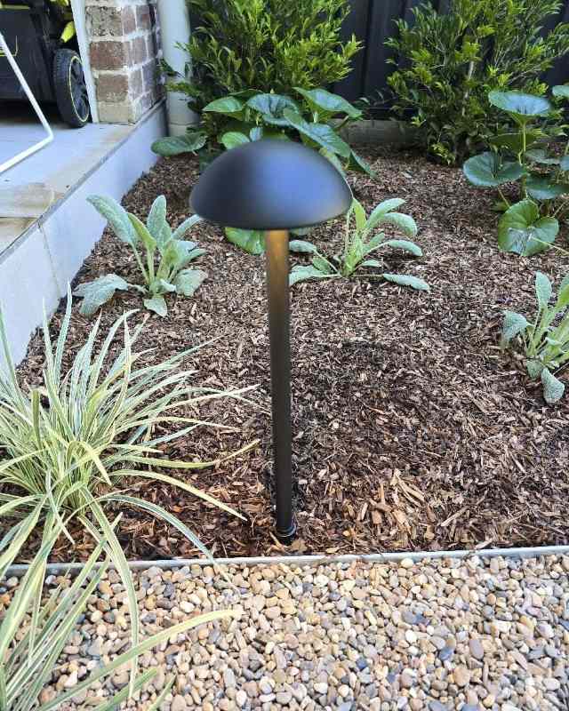 Black mushroom-shaped garden solar light standing in a mulched flower bed with green leafy plants and stone path border.