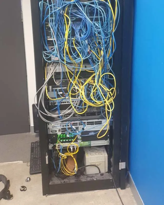 Server rack filled with tangled blue and yellow network cables connected to various networking equipment in a room with a blue wall.